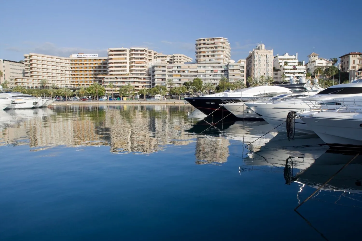 Luxury yachts docked in a calm marina with the Melia Palas Atenea hotel and surrounding buildings in the background.