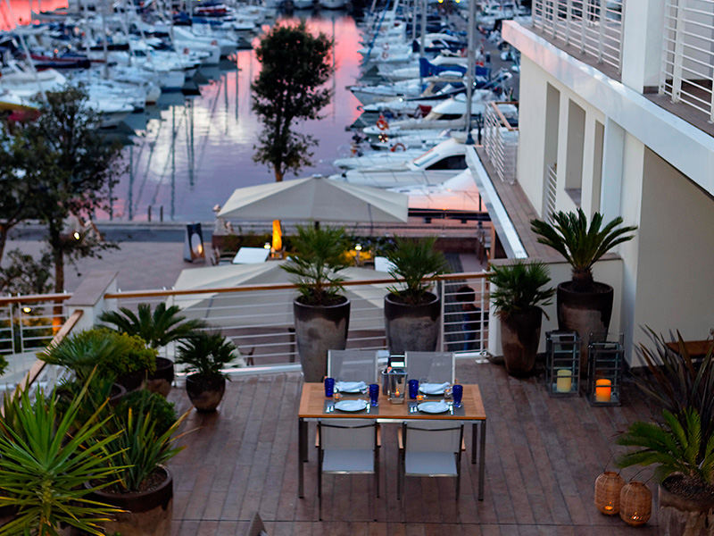 Stylish outdoor dining area on a wooden terrace overlooking a marina at sunset.