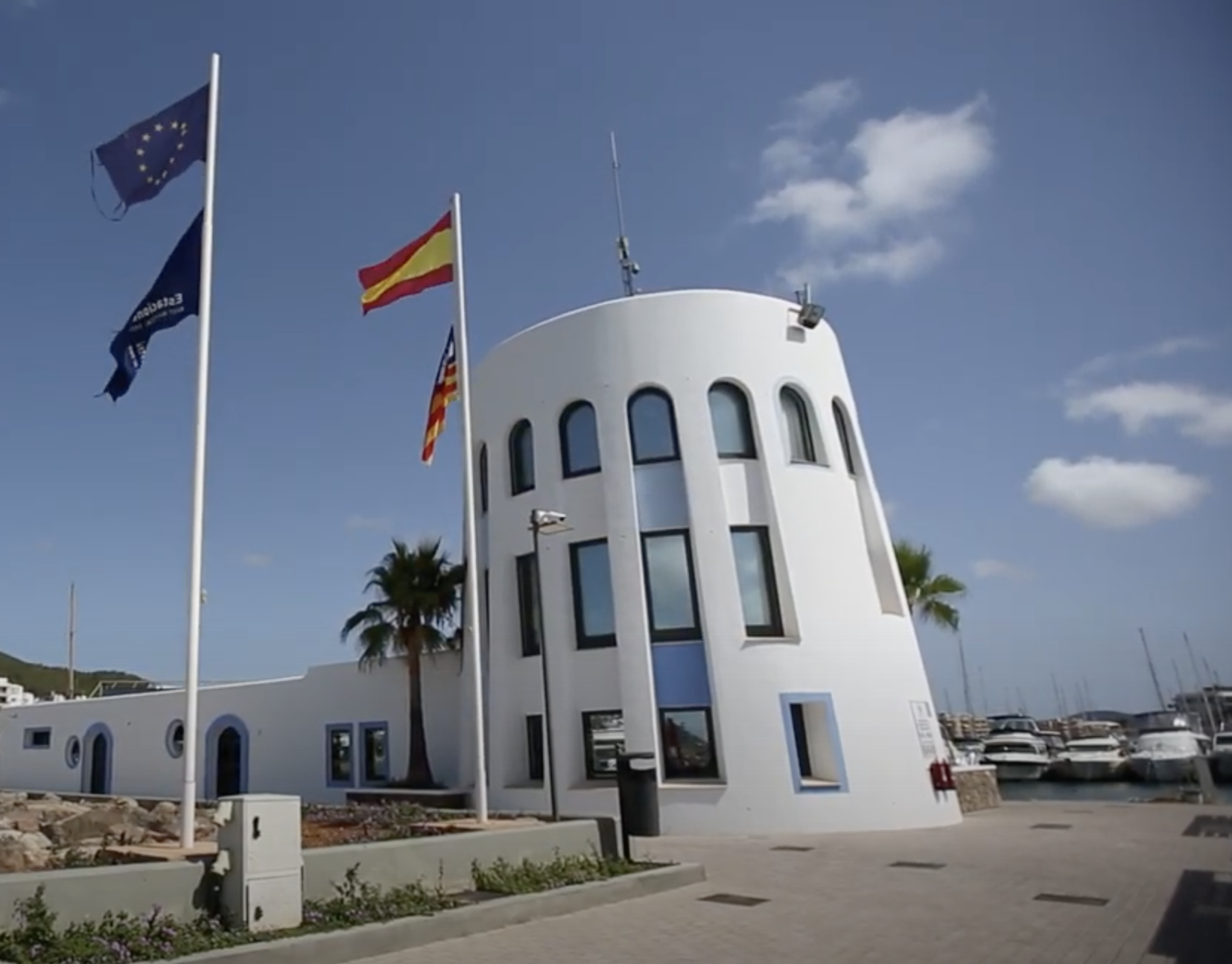 Modern white harbor building with arched windows, palm trees, and three flags in front, located by a marina under a blue sky.