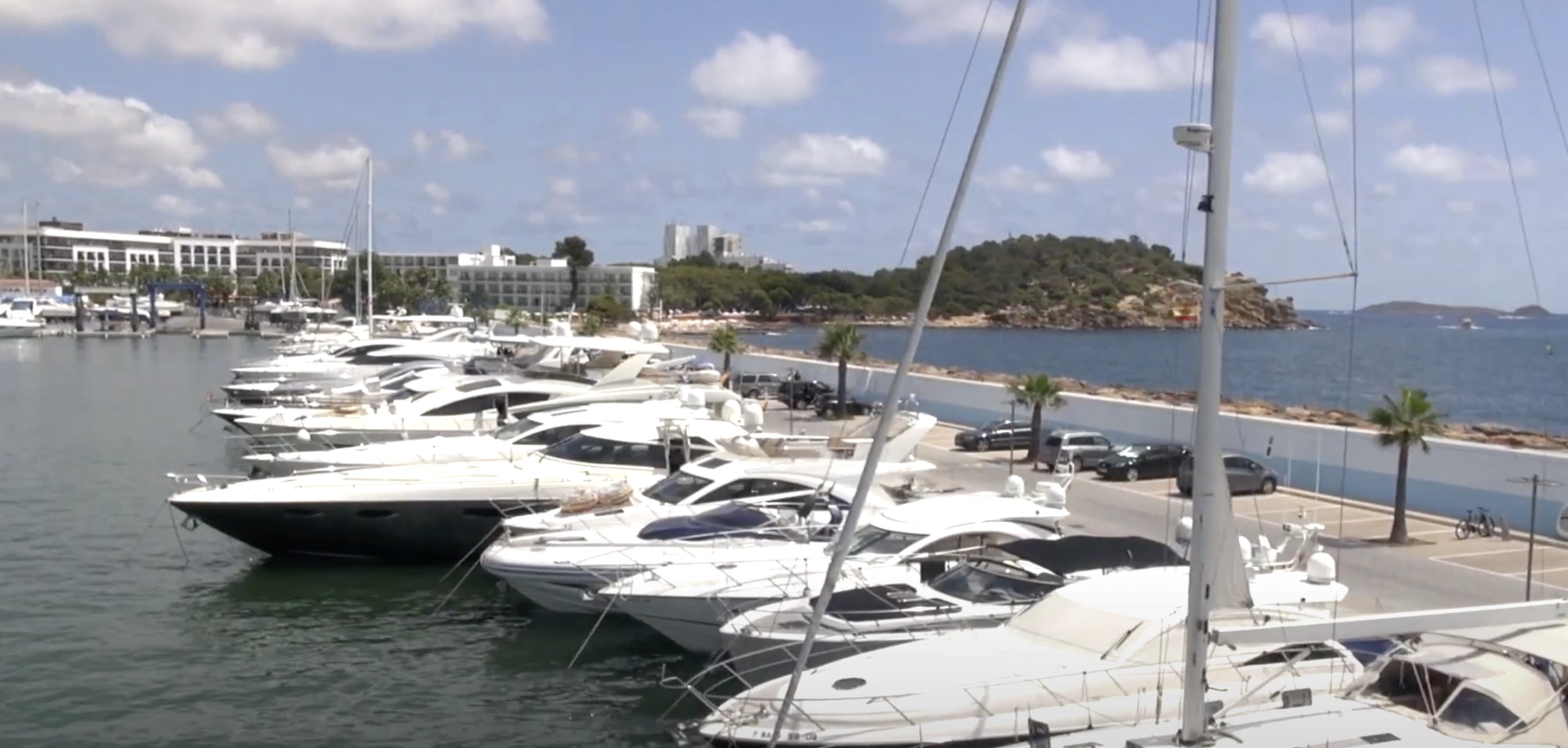 Luxury yachts docked in a sunny marina with a seaside resort and an island in the background.