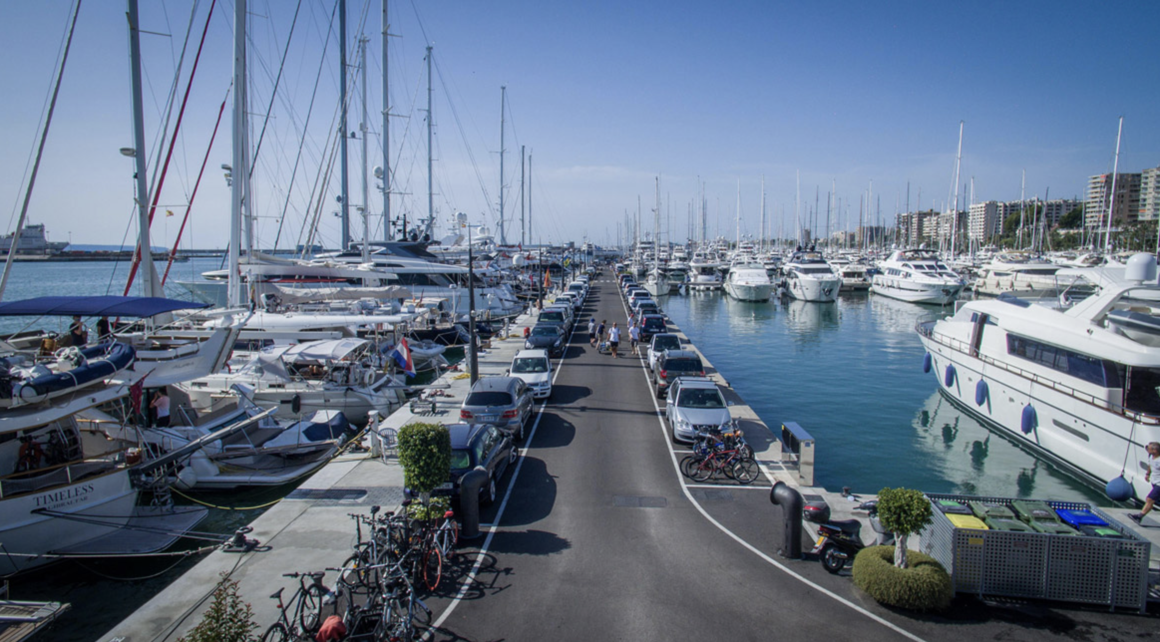 View of a marina filled with yachts and sailboats on both sides of a central pier with parked cars and bicycles.