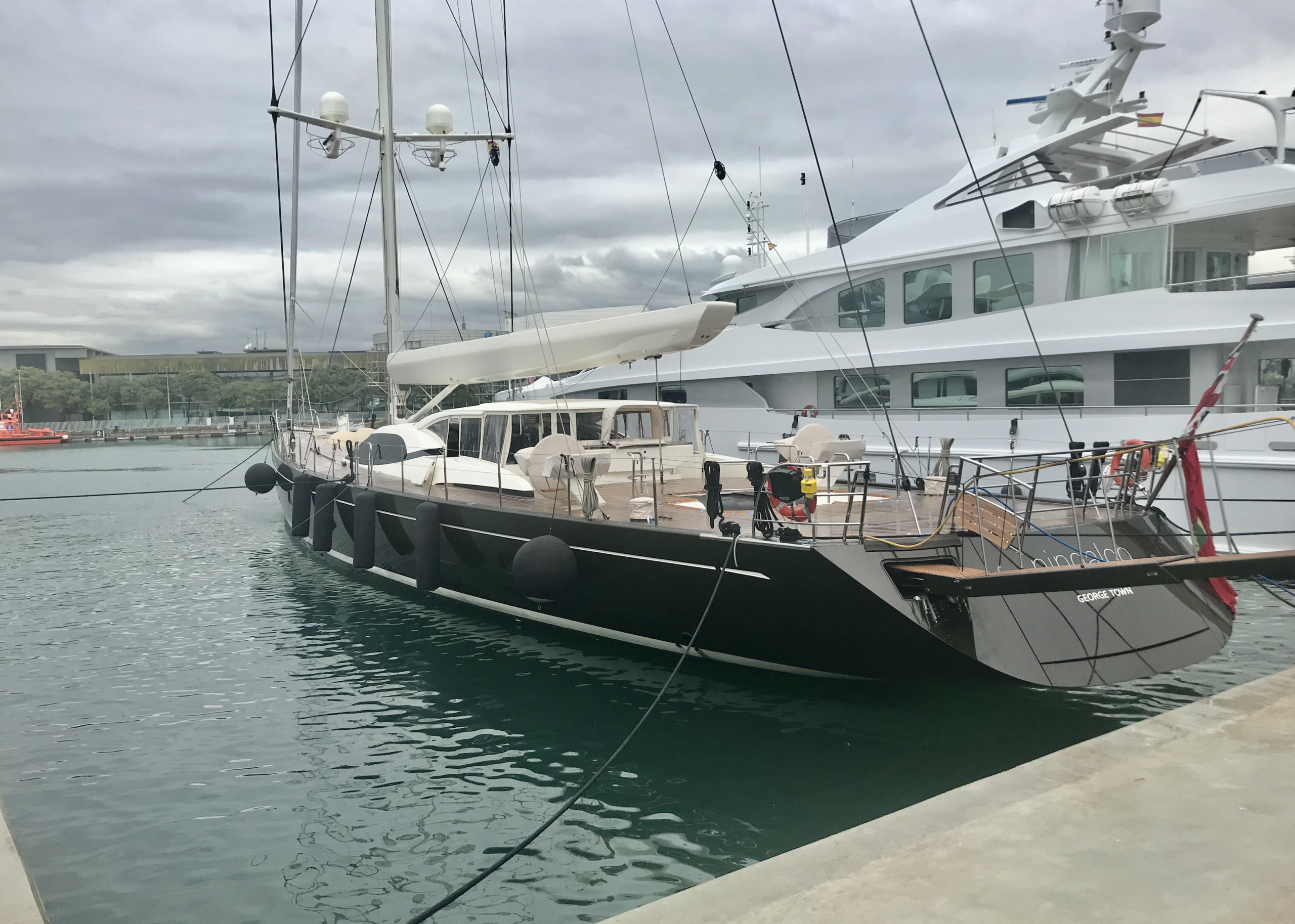 A sleek black sailing yacht named "GIOE I" docked in a marina beside a large white superyacht.