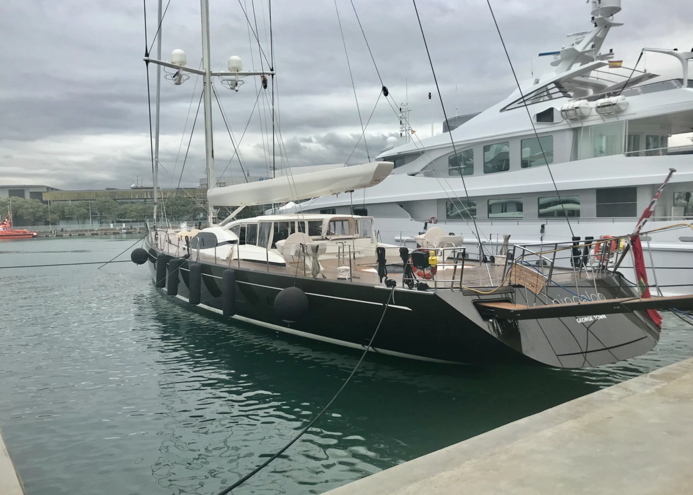 A sleek black sailing yacht named "GIOE I" docked in a marina beside a large white superyacht.