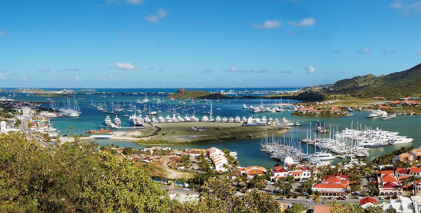 A panoramic view of Simpson Bay Marina in St. Maarten, showcasing numerous yachts, sailboats, and coastal buildings surrounded by turquoise waters and green hills.
