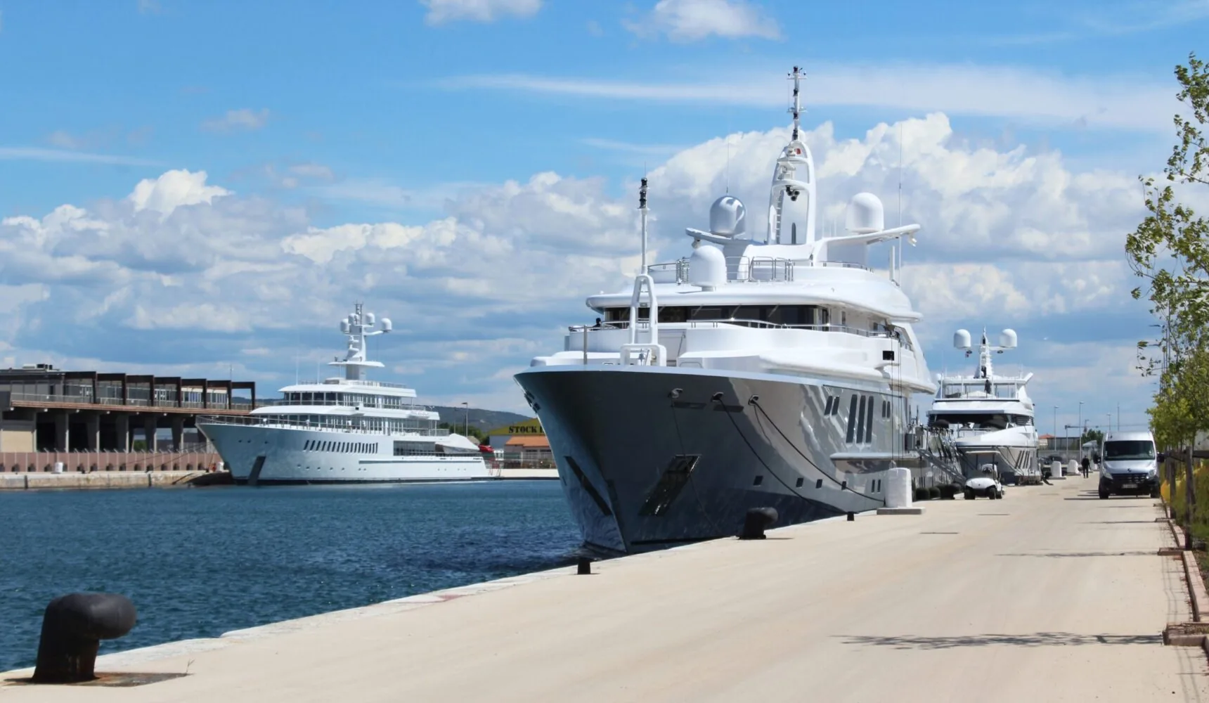 Several luxury yachts, including the superyacht Siren, docked at a sunny marina under a partly cloudy sky.