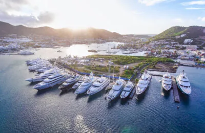 A row of luxurious superyachts docked in a marina with a coastal town and green hills in the background at sunset.