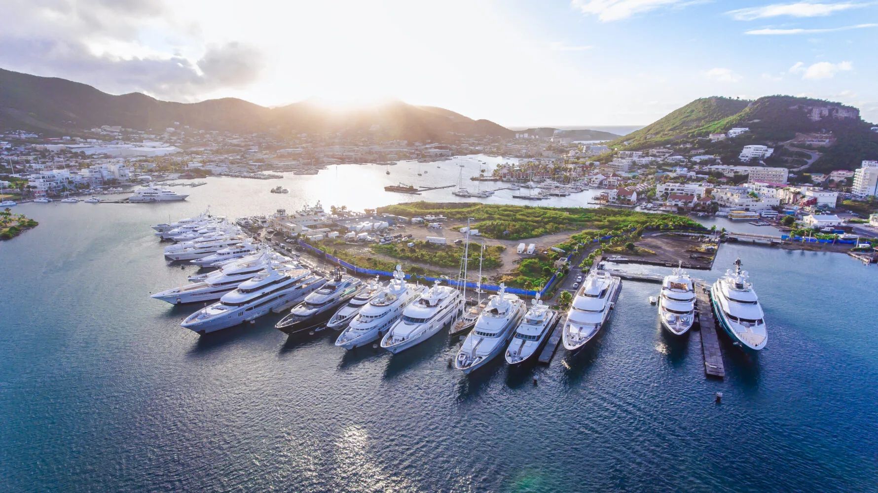 A row of luxurious superyachts docked in a marina with a coastal town and green hills in the background at sunset.