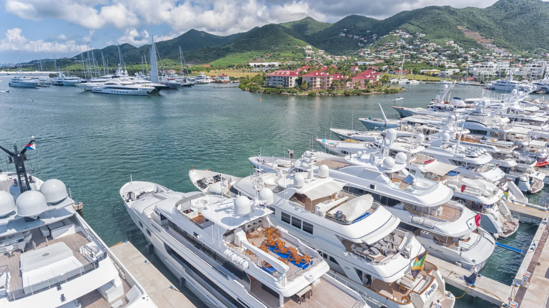 A marina filled with luxury superyachts docked in a tropical bay with green hills and waterfront buildings in the background.