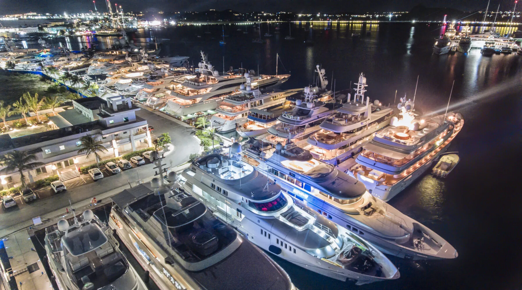 A row of luxury superyachts docked at a brightly lit marina at night.