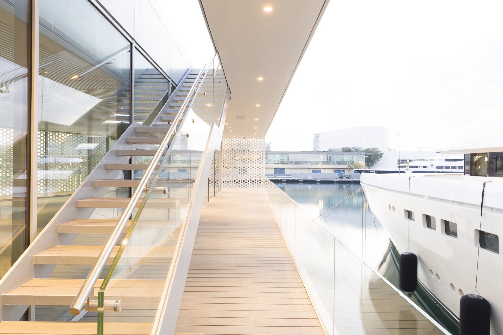 Modern glass walkway of The Gallery Building overlooking a docked luxury yacht in a marina.