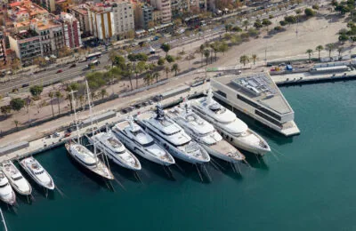 Aerial view of luxury yachts docked at a marina next to The Gallery Building, with a cityscape and palm-lined boulevard in the background.