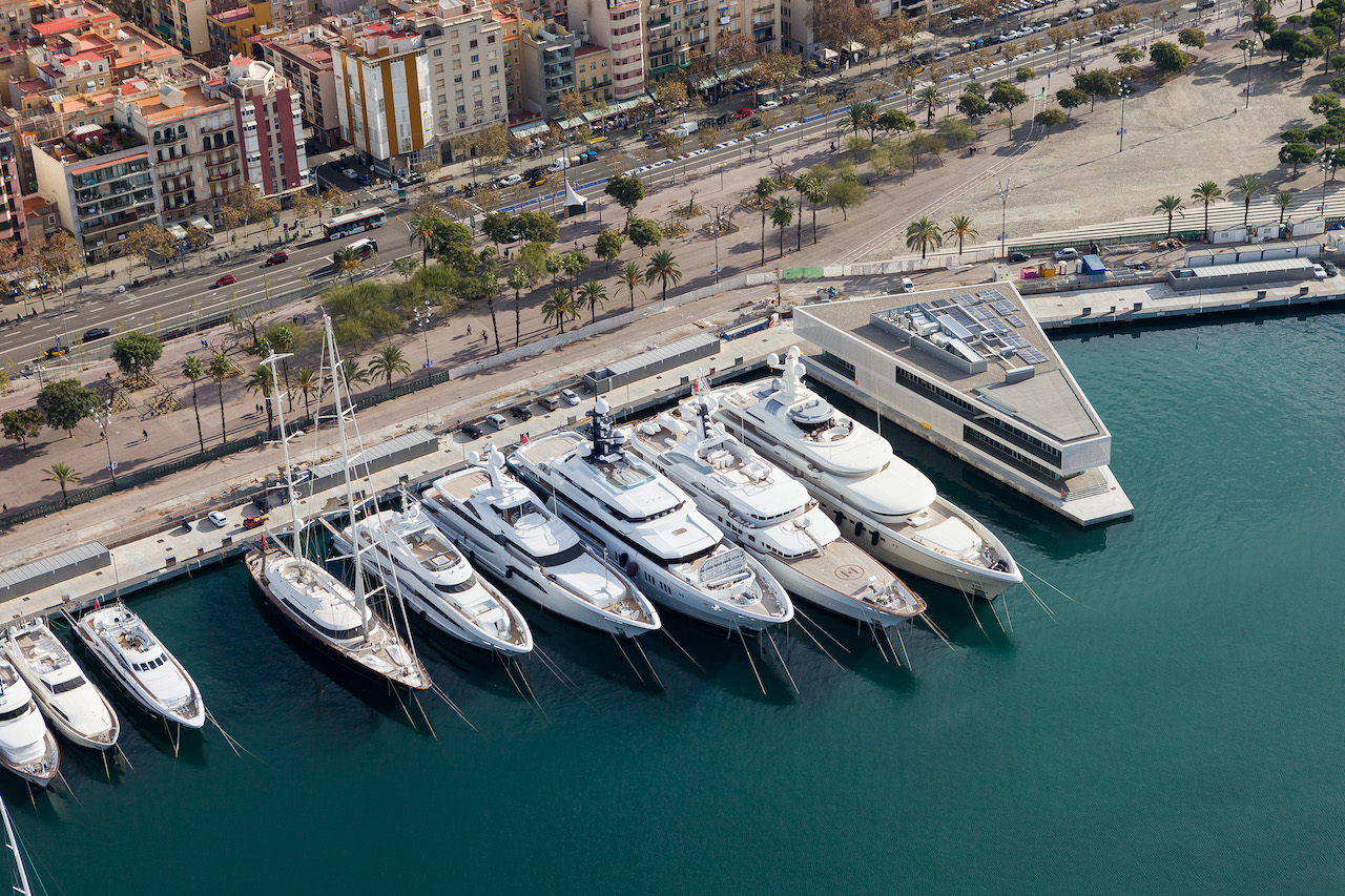 Aerial view of luxury yachts docked at a marina next to The Gallery Building, with a cityscape and palm-lined boulevard in the background.
