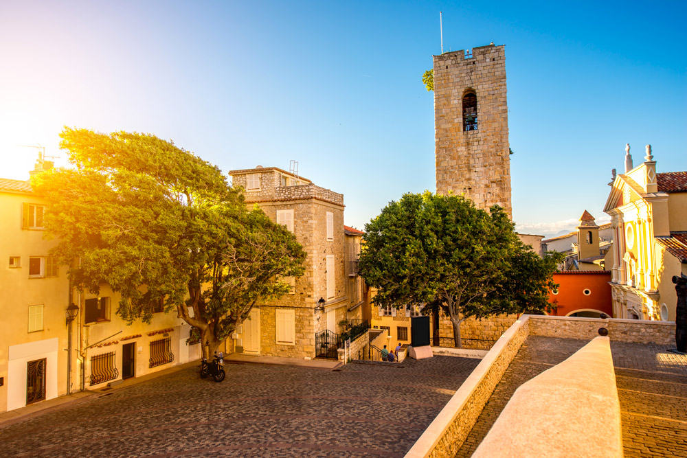 Sunlit view of Place Vauban in Antibes, France, featuring a stone tower, historic buildings, and large trees.
