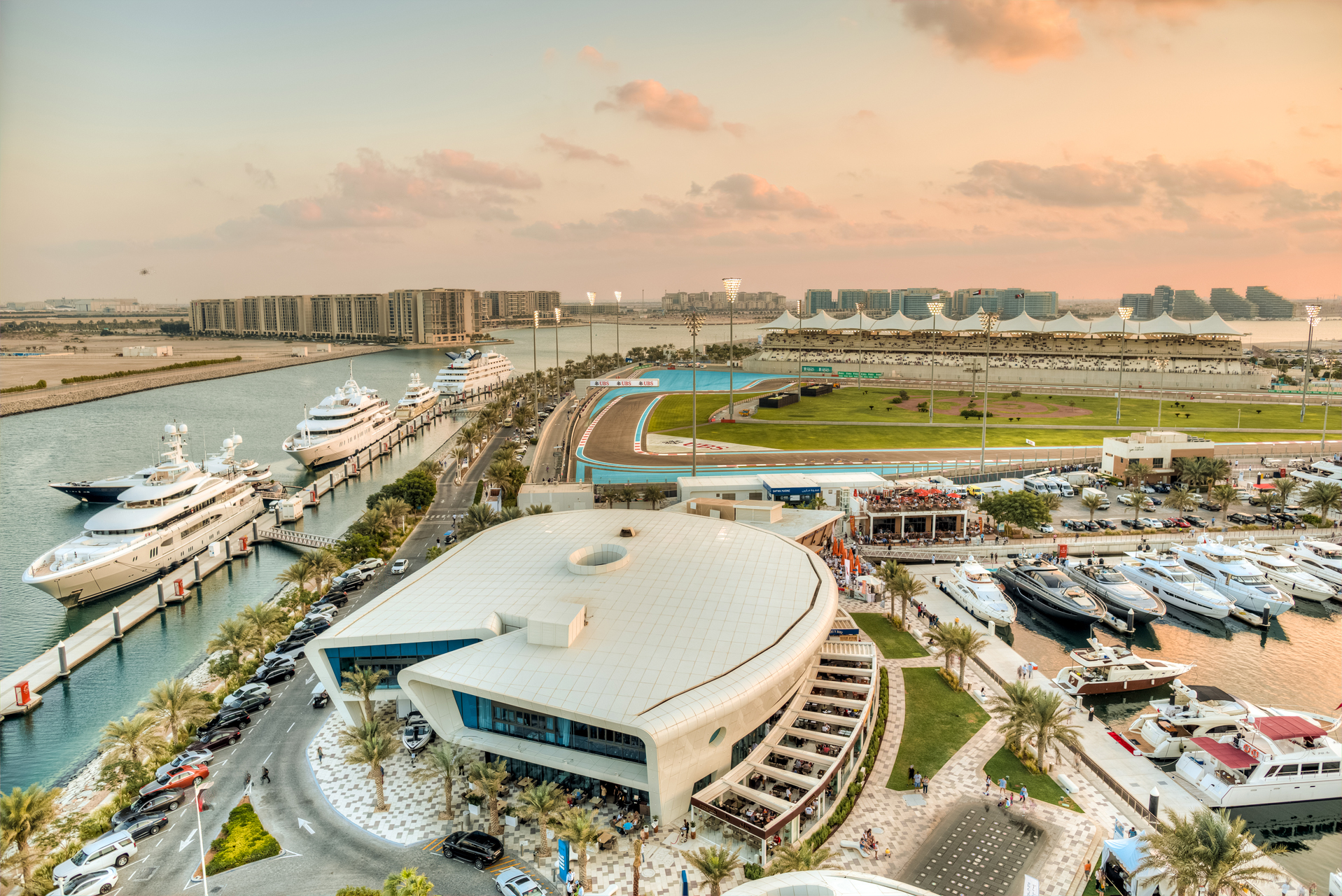 Aerial view of Yas Marina with luxury yachts, modern buildings, and the Yas Marina Circuit racetrack at sunset.