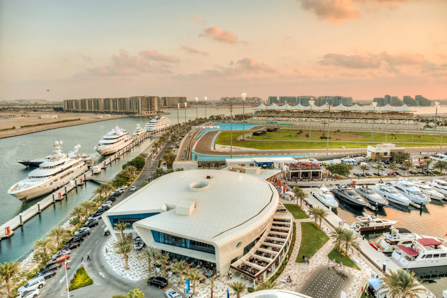 Aerial view of Yas Marina with luxury yachts, modern buildings, and the Yas Marina Circuit racetrack at sunset.