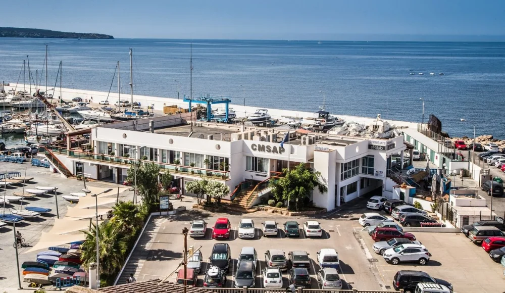 A marina-side building labeled "CMSAP" surrounded by parked cars and sailboats, with a clear view of the sea.