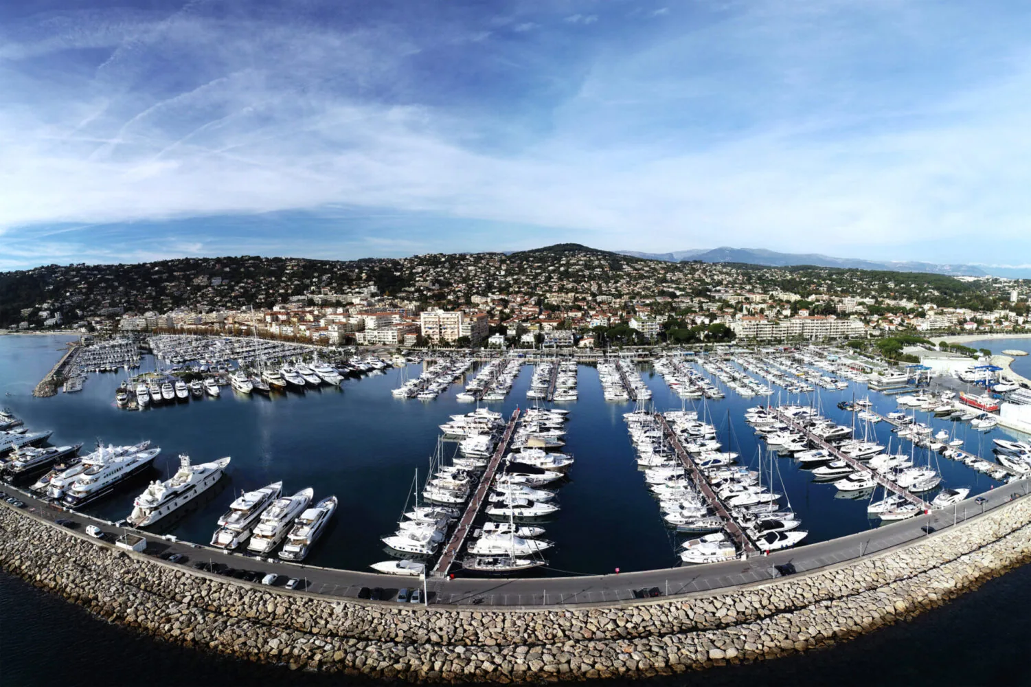 A panoramic view of a marina filled with yachts and boats, with a coastal town and hillside in the background.