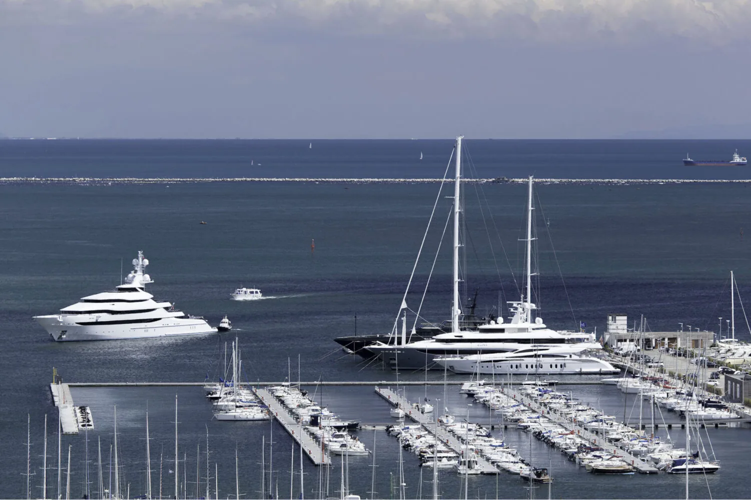 Luxury yachts docked and navigating near a marina in open waters under a cloudy sky.