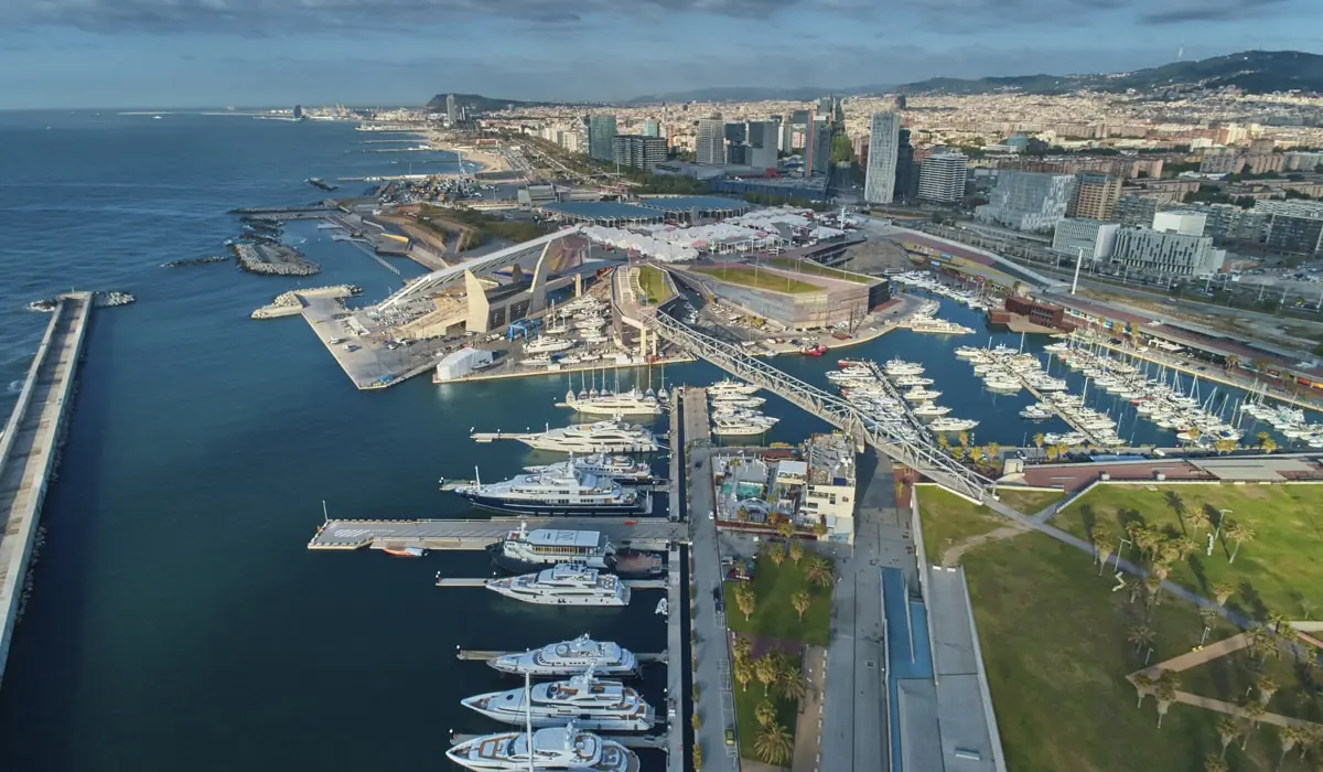 Luftaufnahme des Port Forum in Barcelona mit angedockten Yachten und dem Stadtbild im Hintergrund.