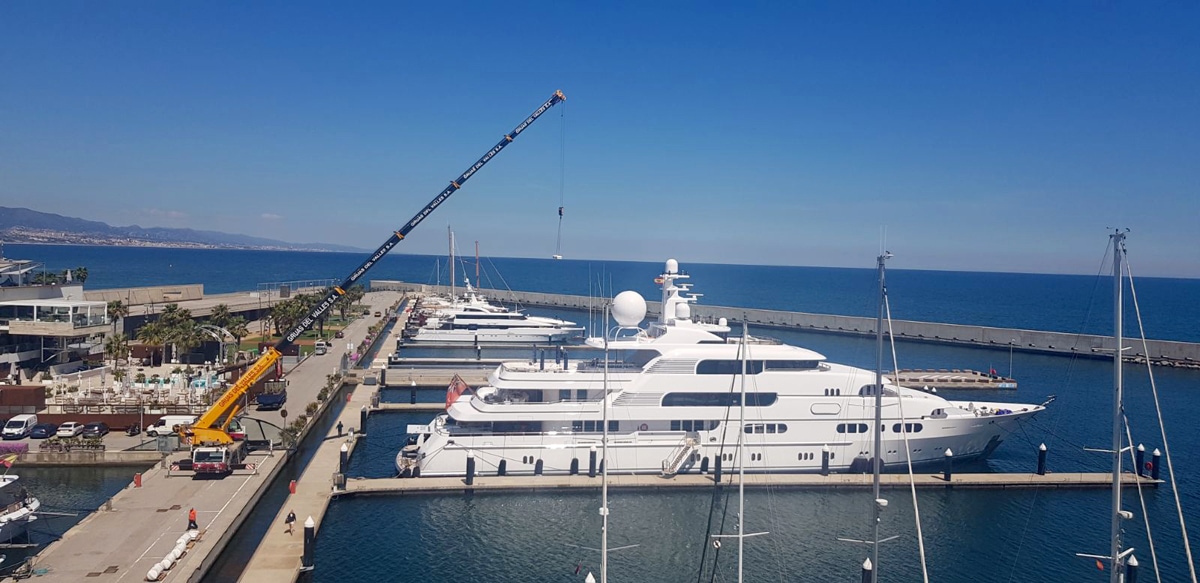 Luxury superyachts docked at Port Fòrum marina with a large crane servicing the pier under a clear blue sky.