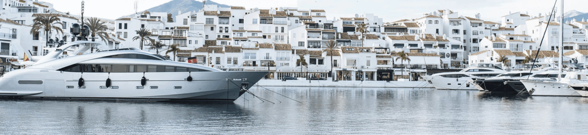 Eine Luxusyacht liegt im Yachthafen von Puerto Banús in Marbella vor Anker, vor dem Hintergrund weißer Gebäude und Palmen.