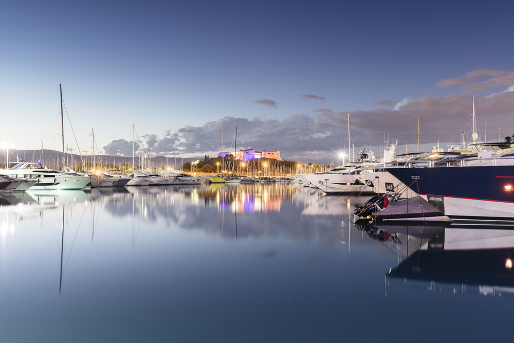 View of Port Vauban in Antibes at dusk, with luxury yachts reflected in the calm water and the illuminated Fort Carré in the background.