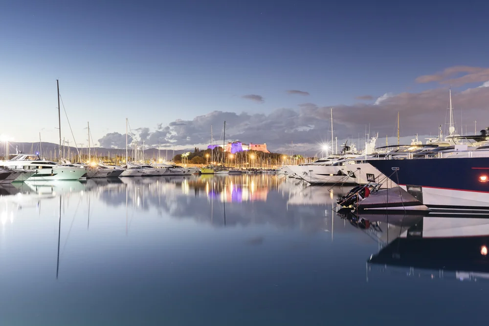 View of Port Vauban in Antibes at dusk, with luxury yachts reflected in the calm water and the illuminated Fort Carré in the background.