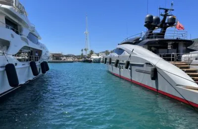 Two luxury yachts docked at a sunny marina with clear blue water and a backdrop of palm trees and hills.