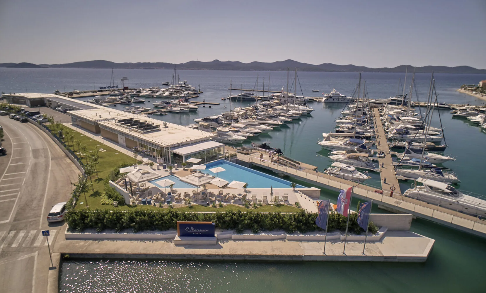 Aerial view of D-Marin Borik marina featuring luxury yachts, a modern waterfront complex, and a poolside lounge area.