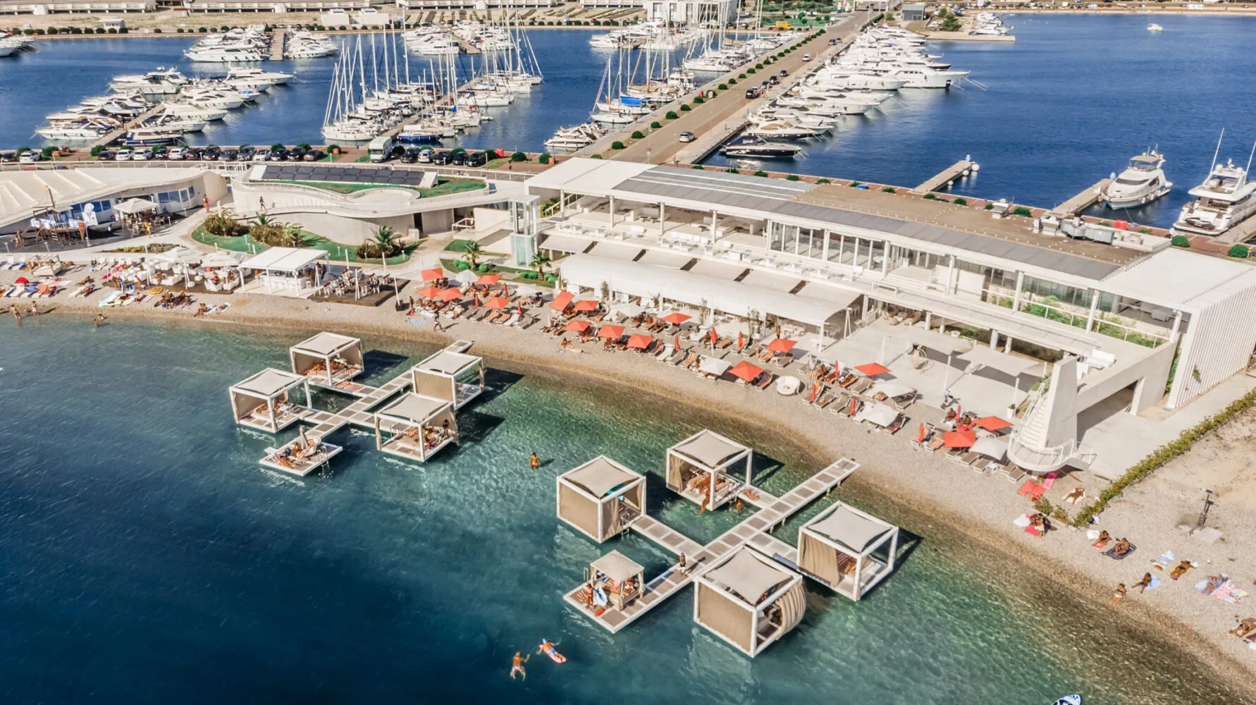Aerial view of D-Marin Dalmacija's Portus Beach Club with floating cabanas, sun loungers, and a marina filled with yachts.