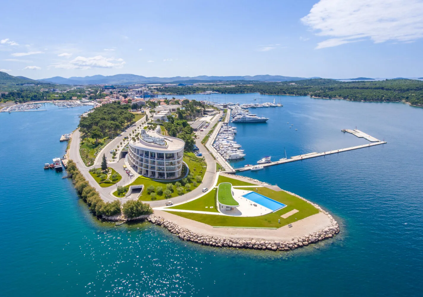 Aerial view of D-Marin Mandalina marina with luxury yachts, modern buildings, and a swimming pool on a scenic peninsula.