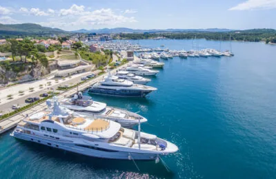 Luxury yachts docked at Mandalina Marina along a scenic coastline with clear blue water and surrounding hills.