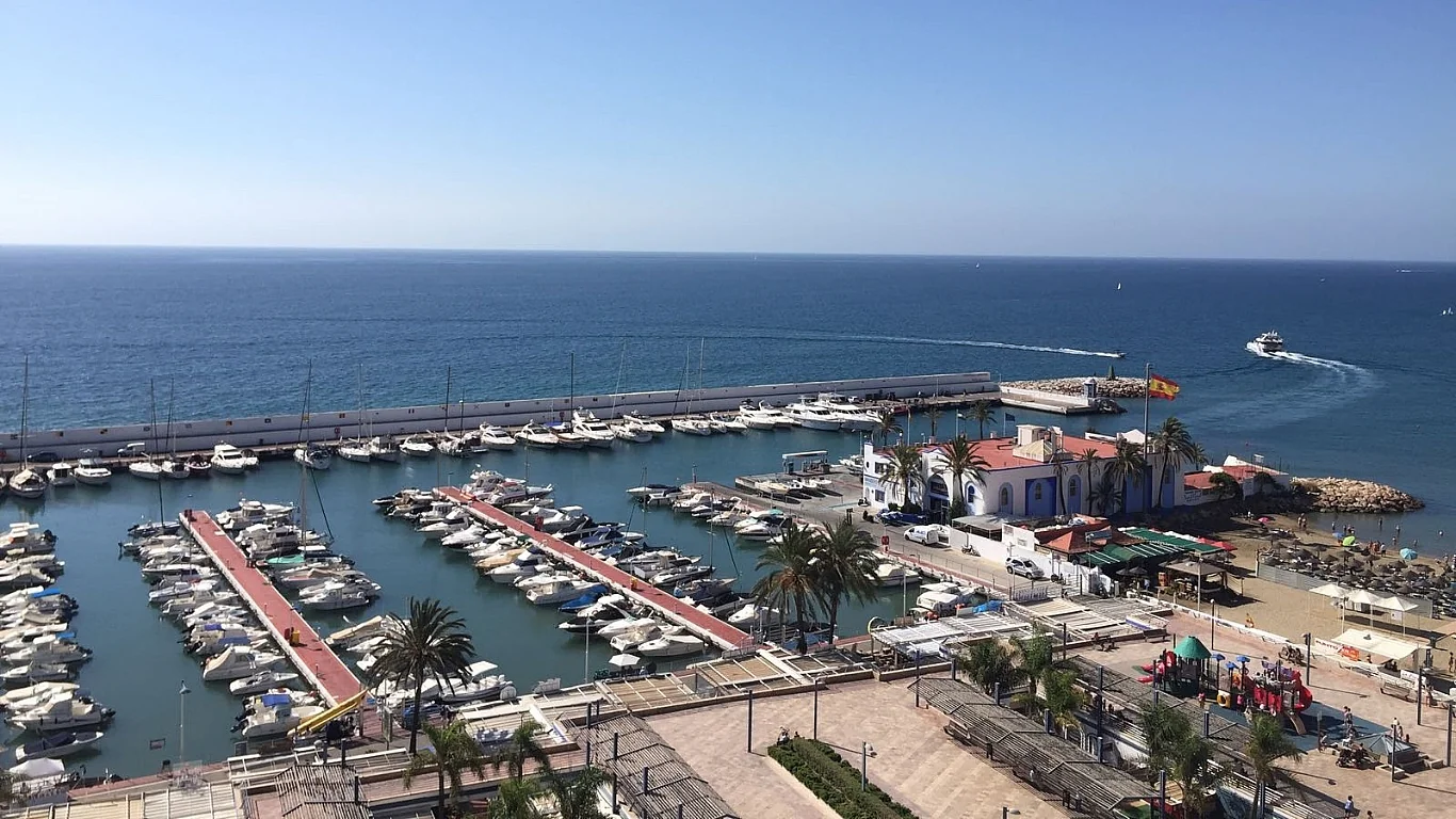 Aerial view of the Puerto Deportivo Marina in Marbella with boats docked, a beach area, and the Mediterranean Sea in the background.