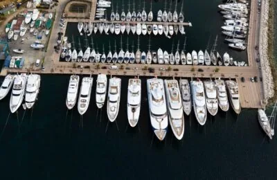 Luftaufnahme eines Jachthafens mit zahlreichen Yachten und Booten, die an einem Pier angedockt sind, mit einer Uferlinie auf der rechten Seite und einem Parkbereich auf der linken Seite.