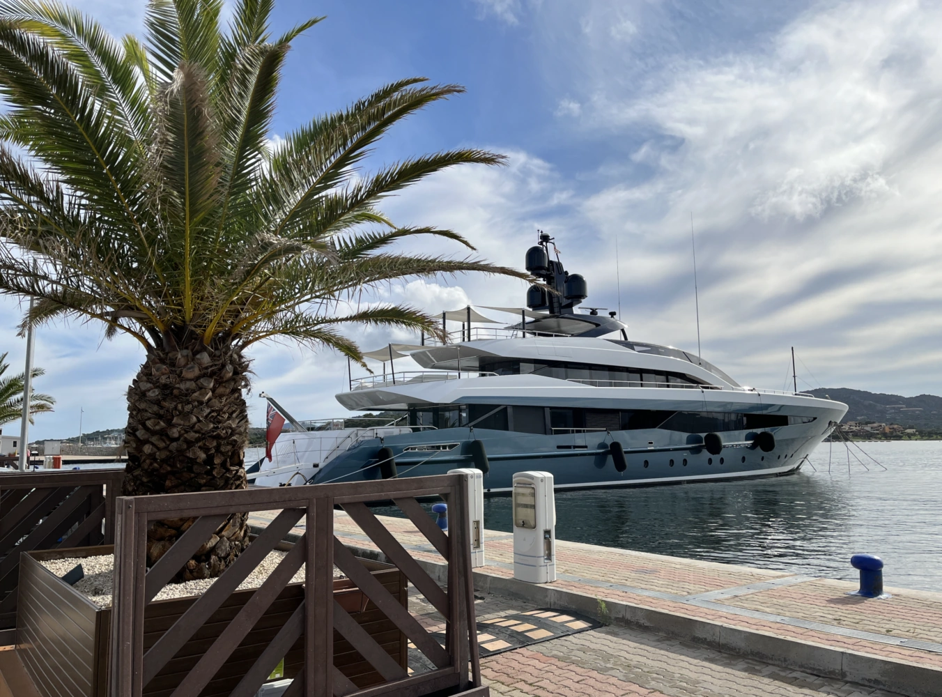 A luxury yacht docked at a harbor with a palm tree in the foreground under a partly cloudy sky.