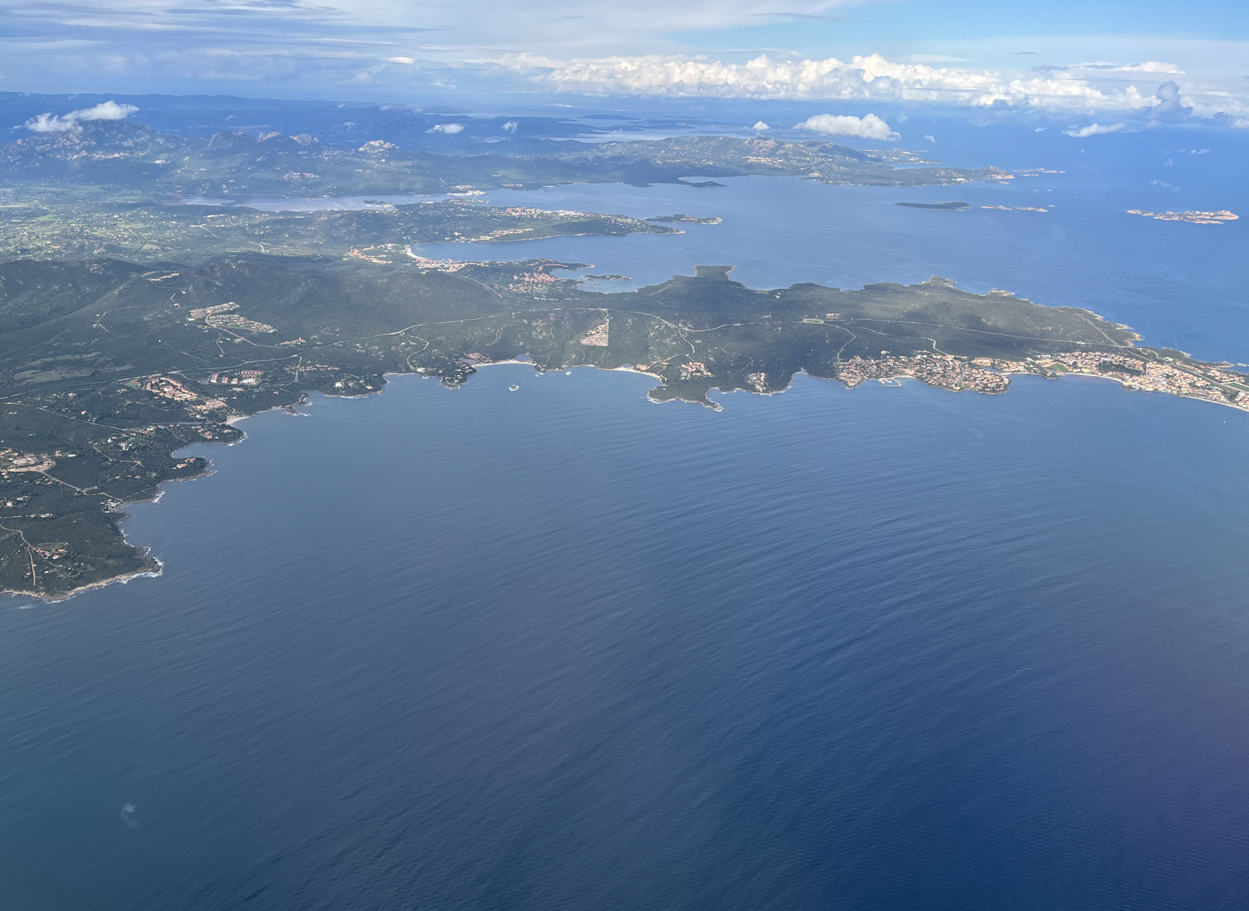 Aerial view of a coastal landscape with lush green hills, winding roads, and deep blue sea near Porto Cervo, Sardinia.