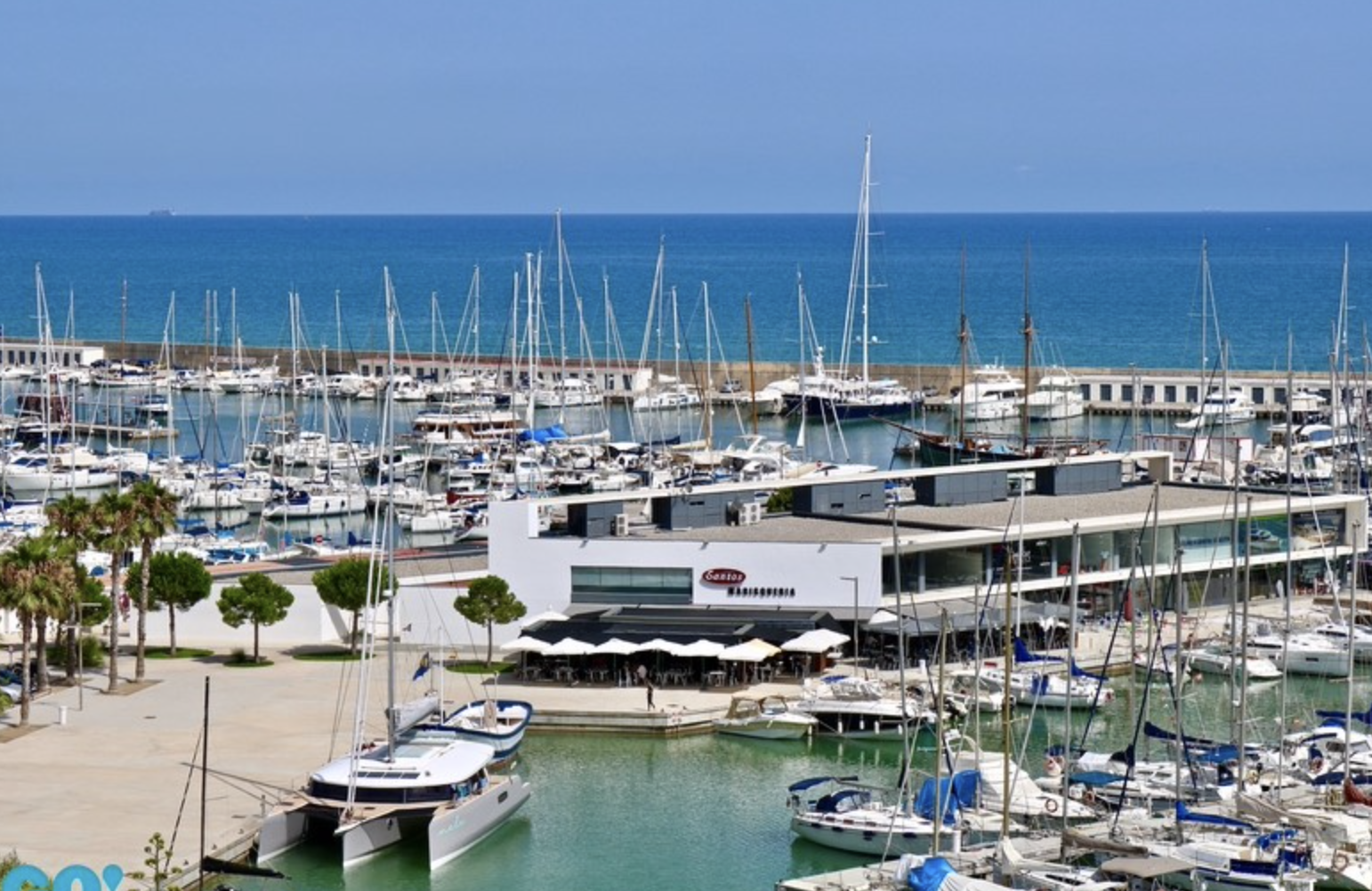 View of a sunny marina filled with sailboats and yachts next to the waterfront restaurant “Sotavento,” with the sea in the background.