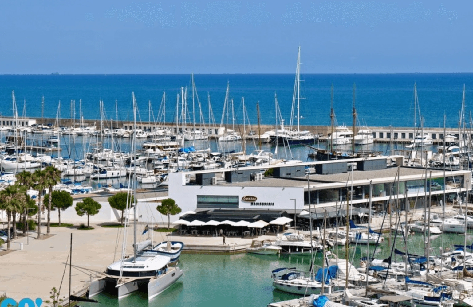 A view of Port Ginesta marina with numerous boats and yachts docked, alongside a modern building and open blue sea in the background.