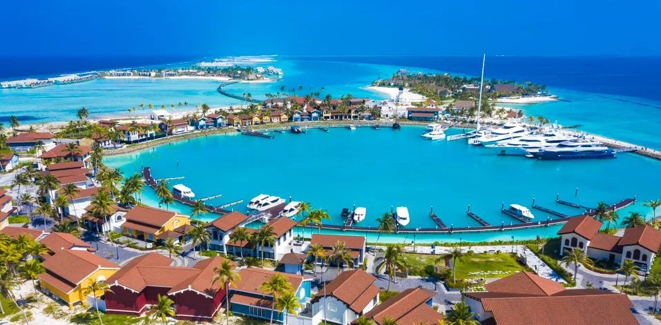 Aerial view of a vibrant yacht marina with turquoise waters, surrounded by palm trees and colorful buildings.