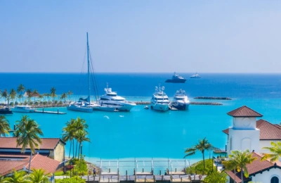 A view of a tropical marina with several luxury yachts anchored in clear blue waters, surrounded by palm trees and a quaint building with a red-tiled roof.