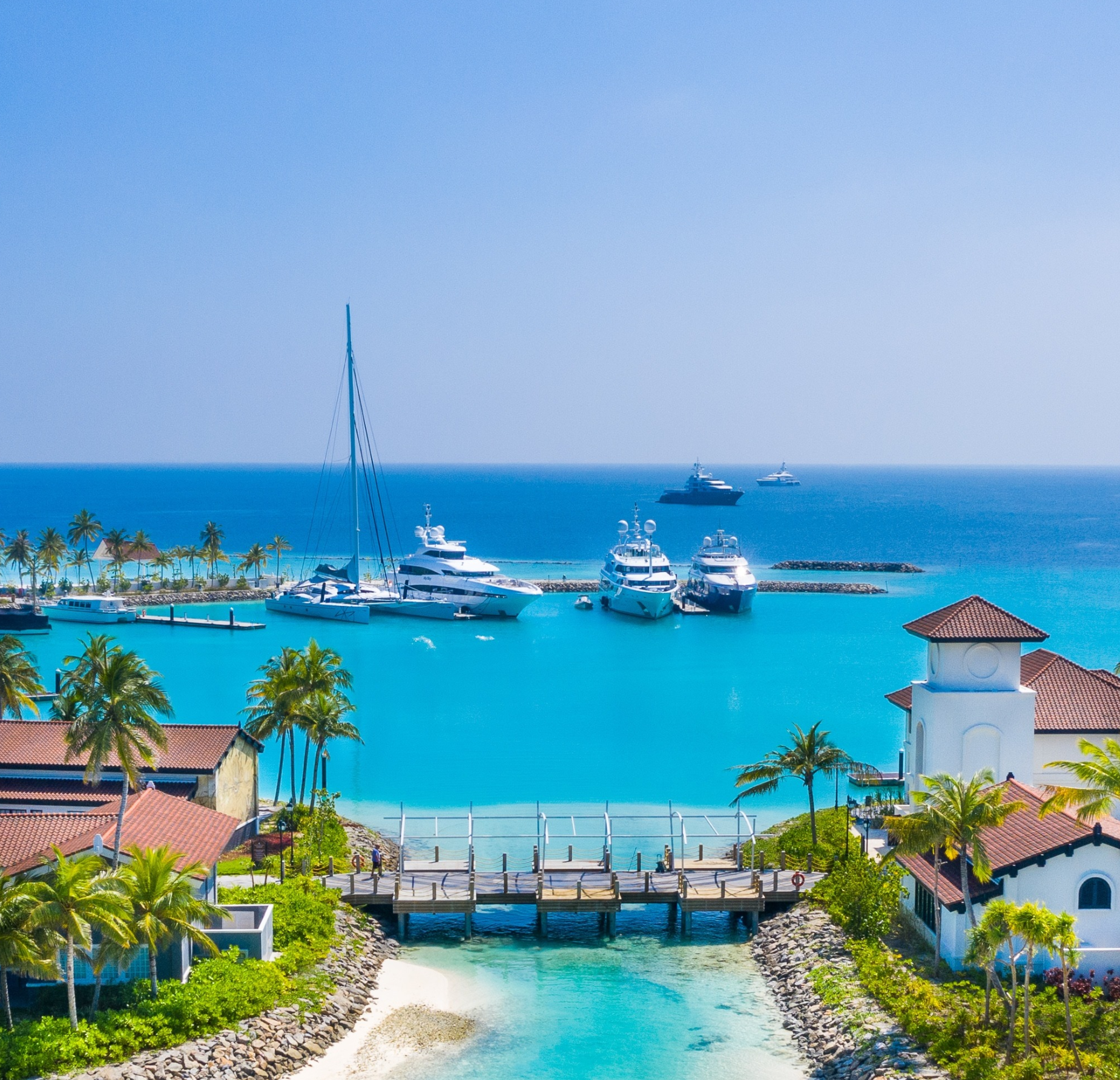 Luxury yachts docked at The Yacht Marina by Jalboot Superyachts in a tropical, turquoise bay surrounded by palm trees and resort buildings.