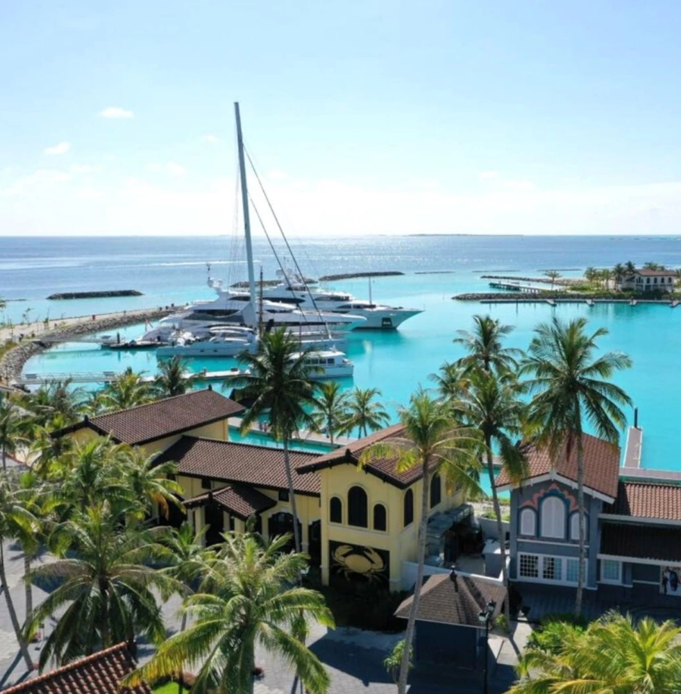 A scenic view of a yacht marina by Jalboot Superyachts, featuring several luxury yachts docked in clear turquoise water surrounded by palm trees and colorful buildings.
