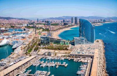 Aerial view of Marina Vela Barcelona, featuring numerous yachts, a prominent sail-shaped hotel, and the city's coastline.