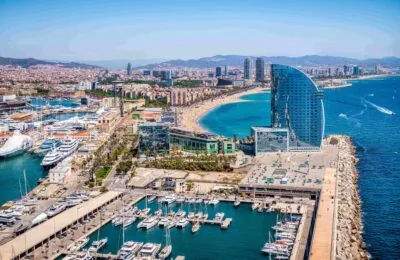 Luftaufnahme des Yachthafens Vela Barcelona mit zahlreichen Yachten, einem markanten Hotel in Form eines Segels und der Küstenlinie der Stadt.