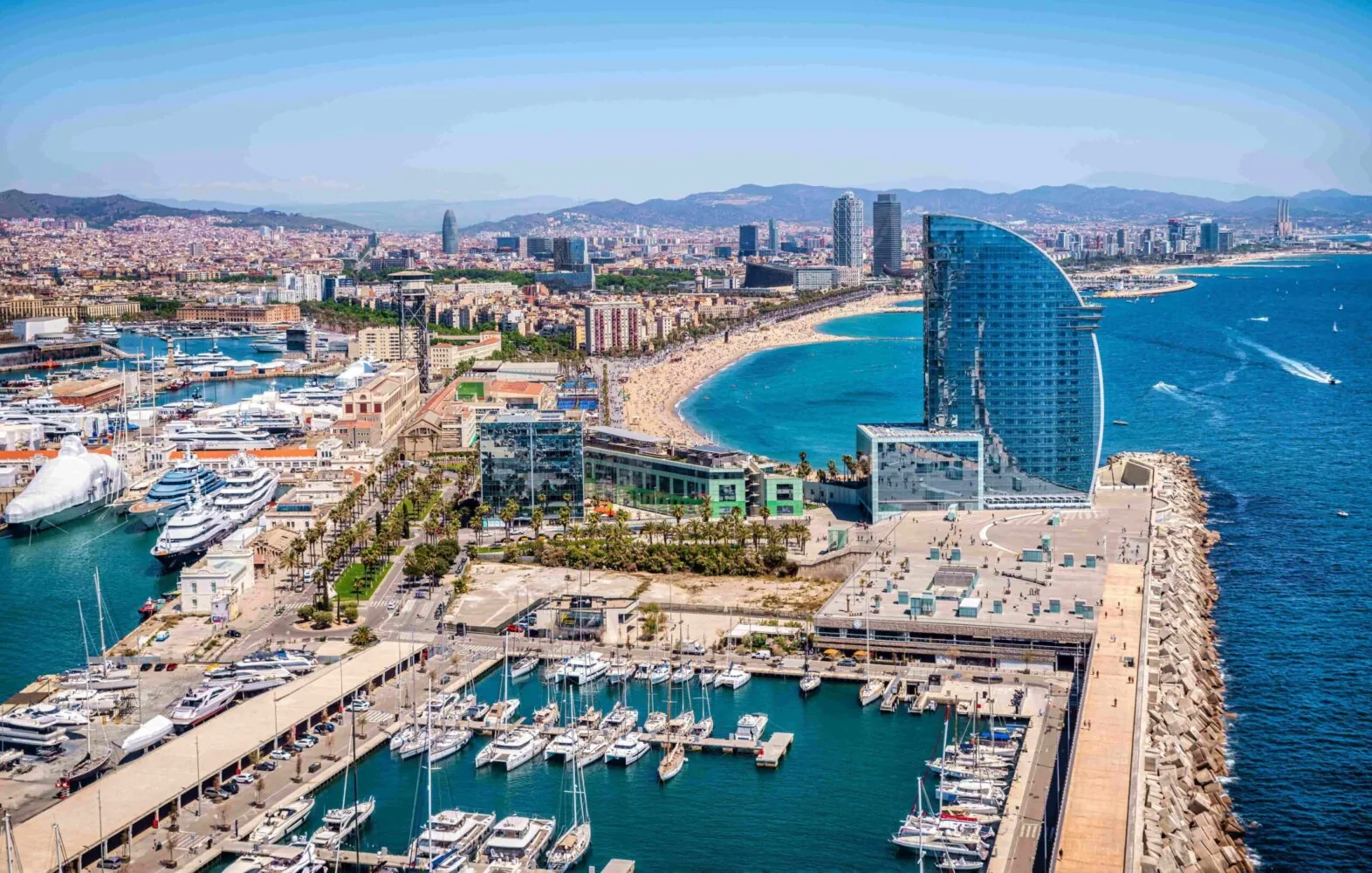 Aerial view of Marina Vela Barcelona, featuring numerous yachts, a prominent sail-shaped hotel, and the city's coastline.