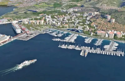 Aerial view of a coastal city with cruise ships, a large marina full of yachts, and urban buildings surrounded by green hills.