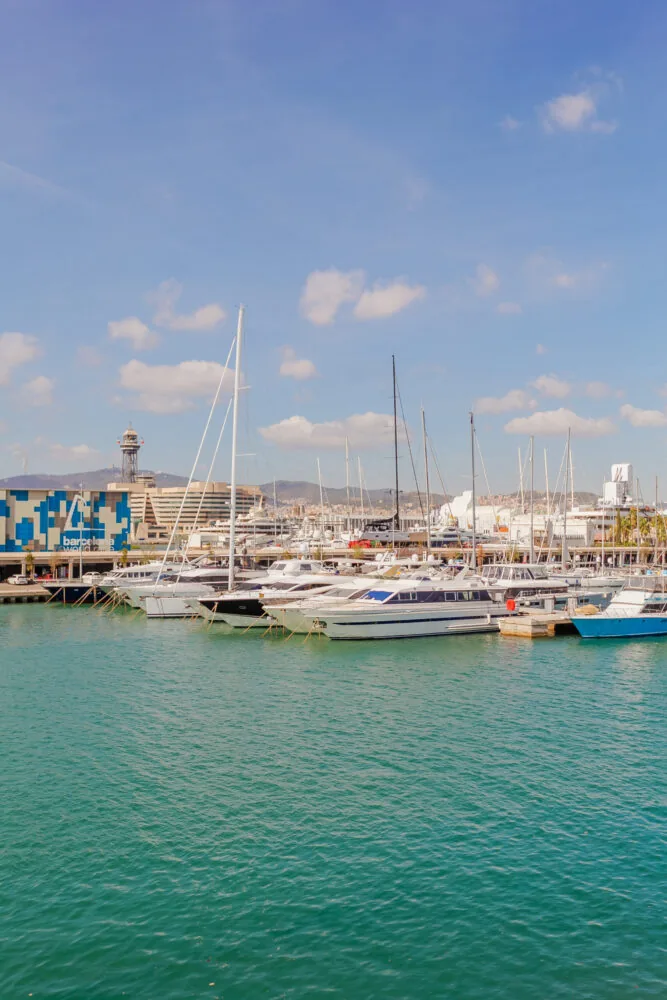 A marina with moored yachts and sailboats on a sunny day in Barcelona, with a cityscape and blue sky in the background.