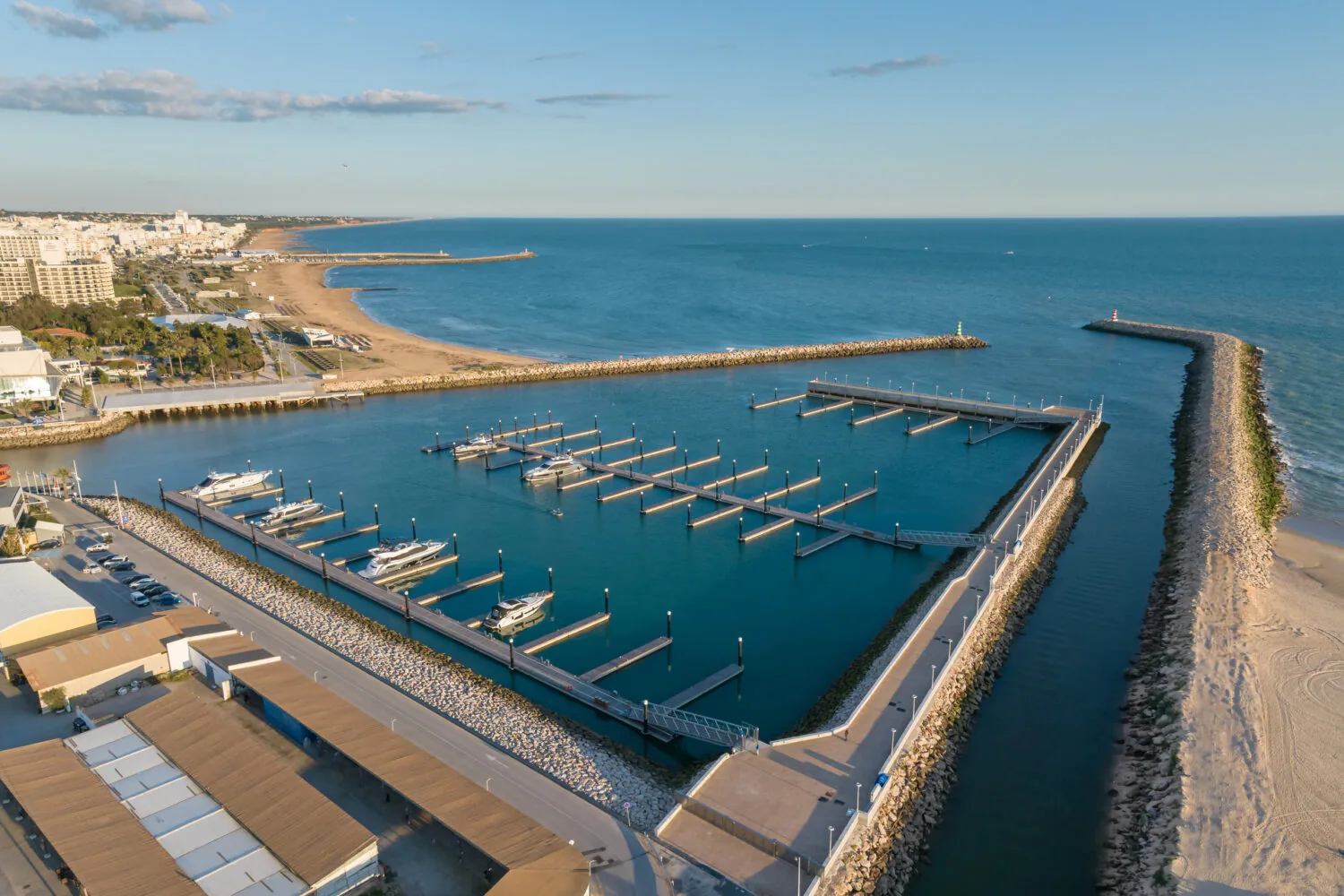 Aerial view of VLM Marina featuring multiple docks and moored yachts, with a sandy beach and town in the background.
