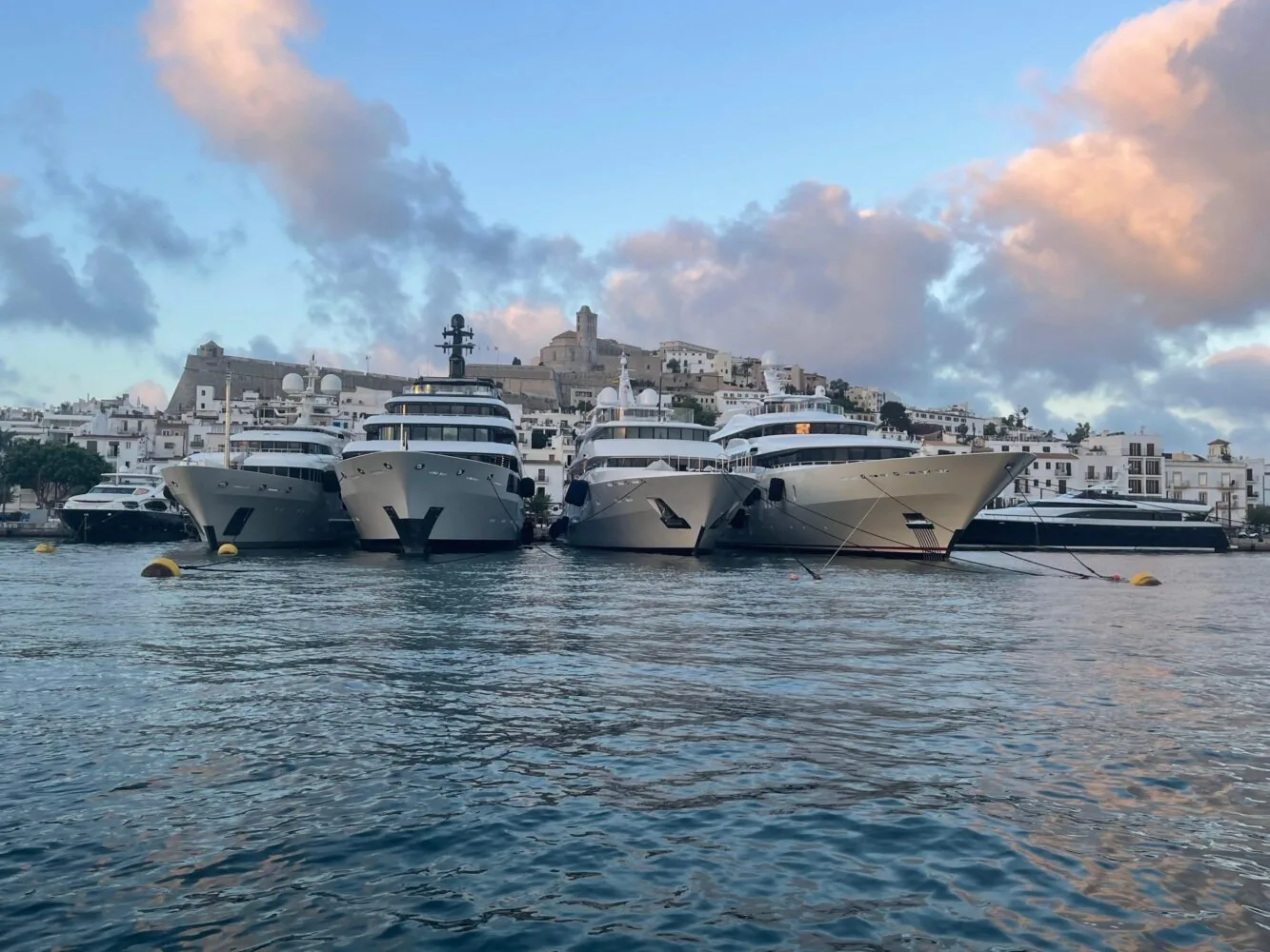 Four large yachts are moored side by side in Marina Ibiza with a picturesque hillside town in the background under a sky with colorful clouds.