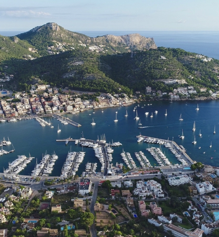 Luftaufnahme von Port Andratx mit zahlreichen vor Anker liegenden Segelbooten, umgeben von üppig grünen Hügeln, wobei der Yachthafen und die Architektur der Küste besonders hervorgehoben werden.